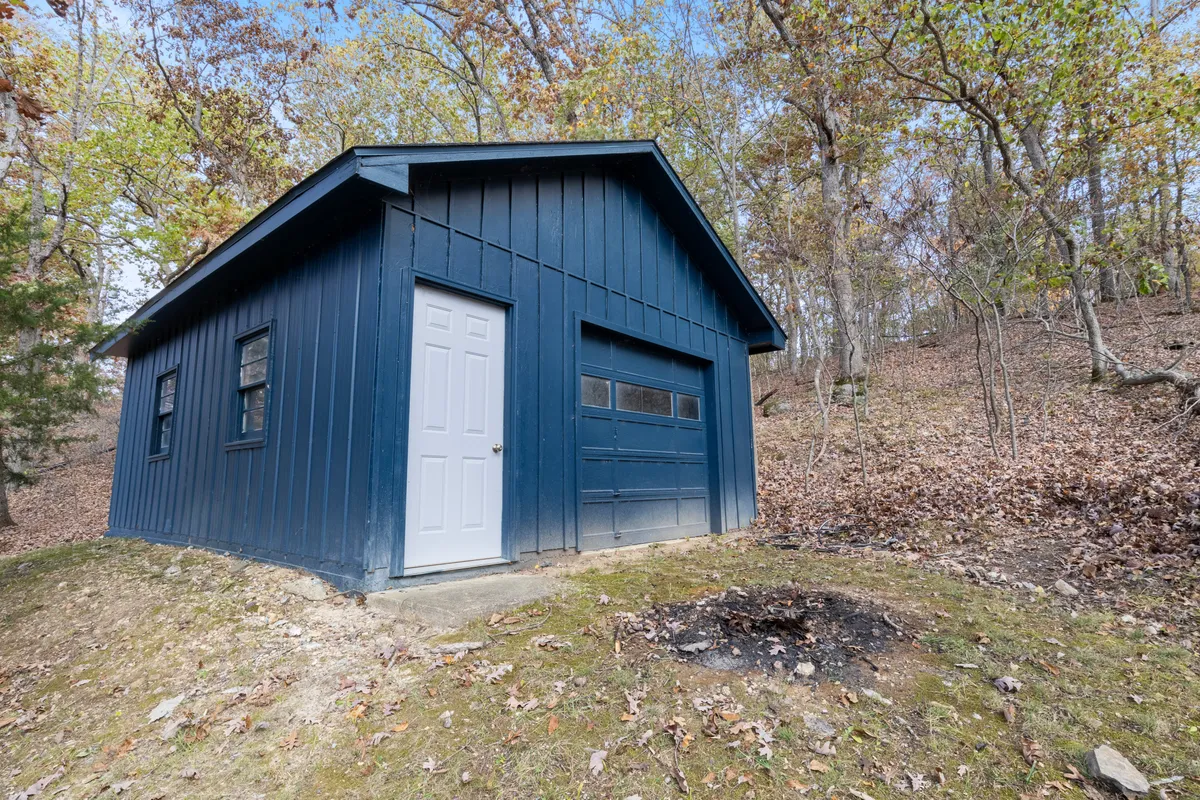A blue shed sits beside a white door, surrounded by lush trees and featuring a fire pit in the foreground. The area invites guests to gather for cozy evenings or seasonal get-togethers, set against a backdrop of greenery that changes with the seasons.