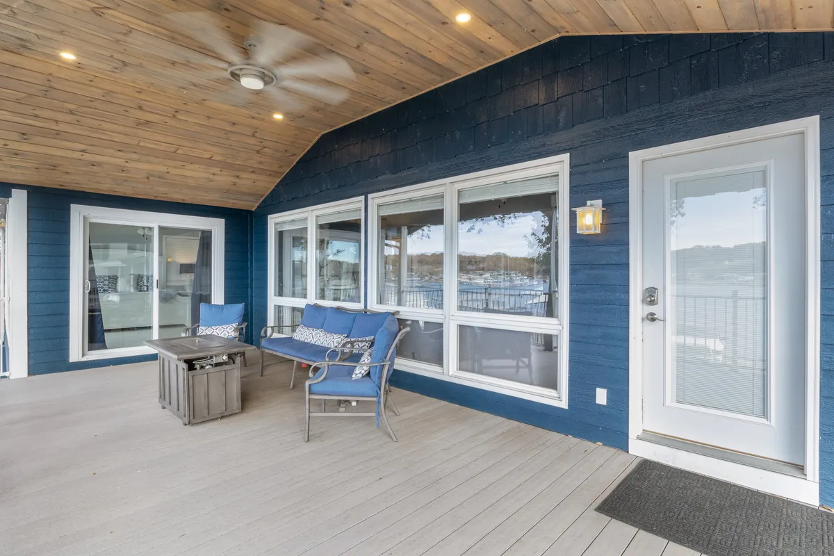 An inviting exterior shot highlights the blue exterior walls, large windows, and a comfortable outdoor seating area beneath a wooden ceiling, complete with a lantern-style light fixture. The ambiance is tranquil, ideal for outdoor dining or simply relaxing while pets rest nearby.