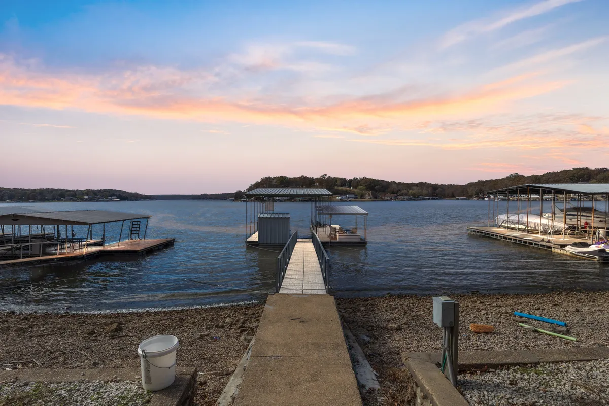 A tranquil dock extends over the serene lake, with boats moored and the expansive water reflecting a soft sky. The scene captures the peaceful allure of lakeside activity, with ample space for watercraft and direct access to the water for swimming, fishing, or embarking on boating adventures.