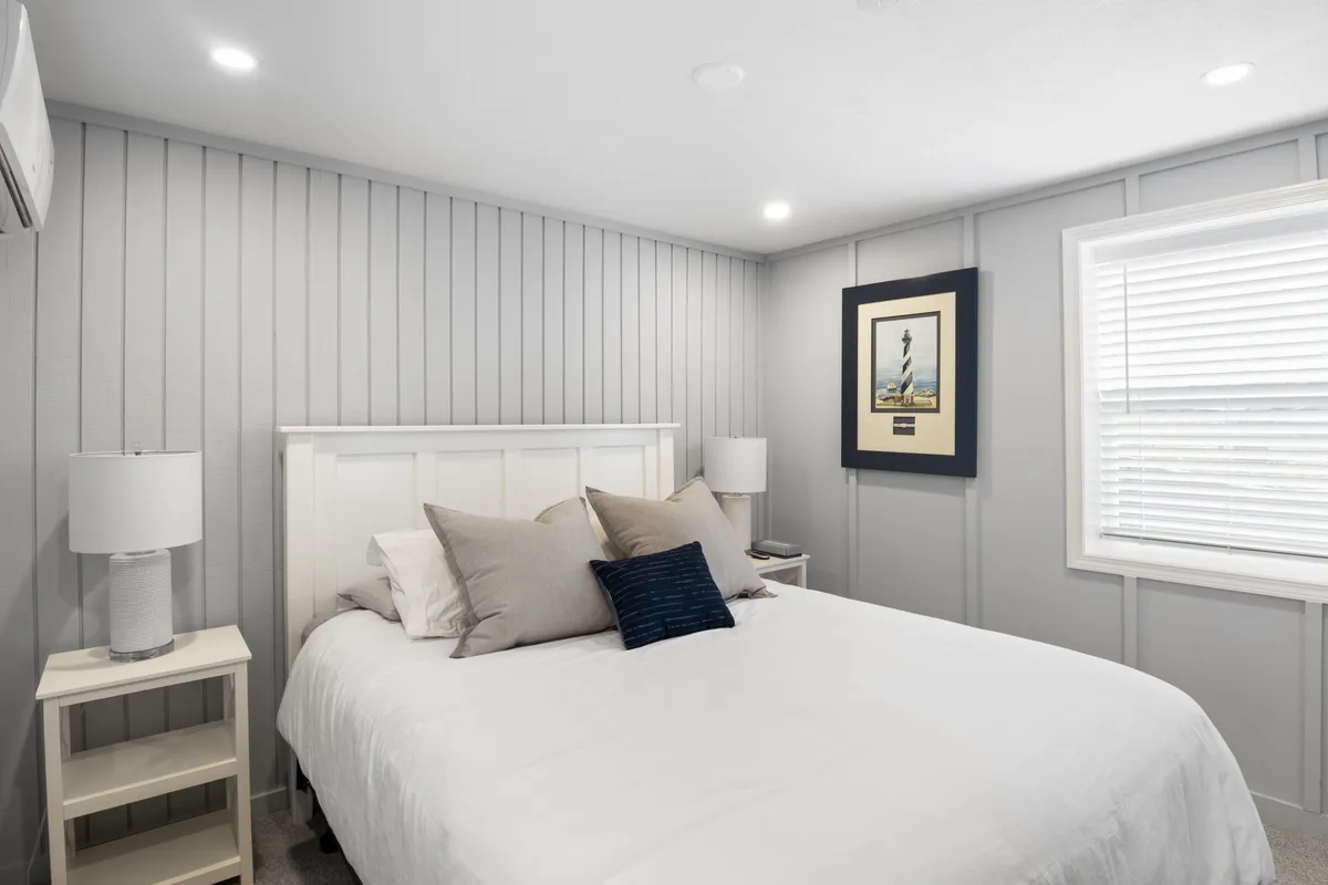 A simple, serene bedroom with a bed, framed artwork above, and a nightstand with a lamp. The soft morning light and uncluttered decor contribute to a peaceful environment ideal for unwinding.