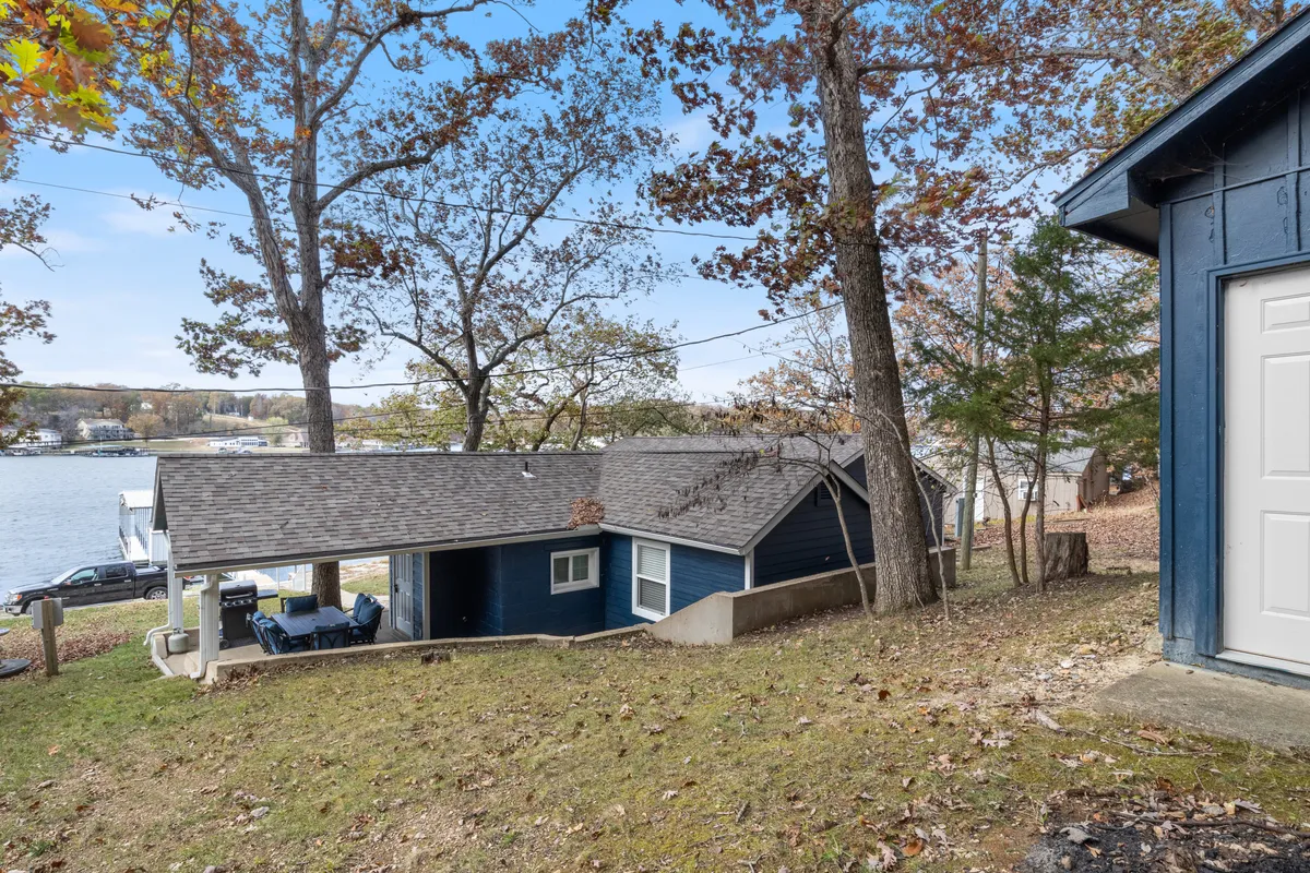 This exterior image features the blue house with a spacious outdoor seating area overlooking the lake, set among mature trees. The setting is tranquil, with plenty of open space for pets to roam on a leash and families to relax together while enjoying the scenic lake views.