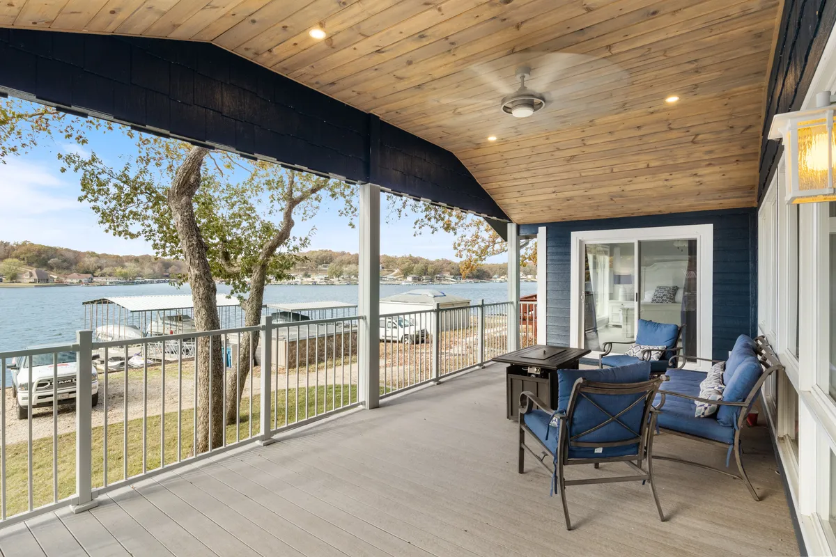 An inviting outdoor seating area on a covered deck overlooks the lake. The space features ceiling fans and wooden ceilings, with comfortable seating arranged to take in the tranquil water view. The scene evokes the pleasures of relaxing outdoors, whether with a morning coffee, a book, or during an evening gathering.