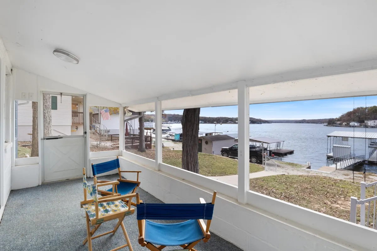 A serene lakefront view from the outdoor seating area, with the entrance door visible. The peaceful water, comfortable seating, and lush greenery create a sense of tranquility and connection to nature. The setting invites guests to relax and enjoy the outdoors.