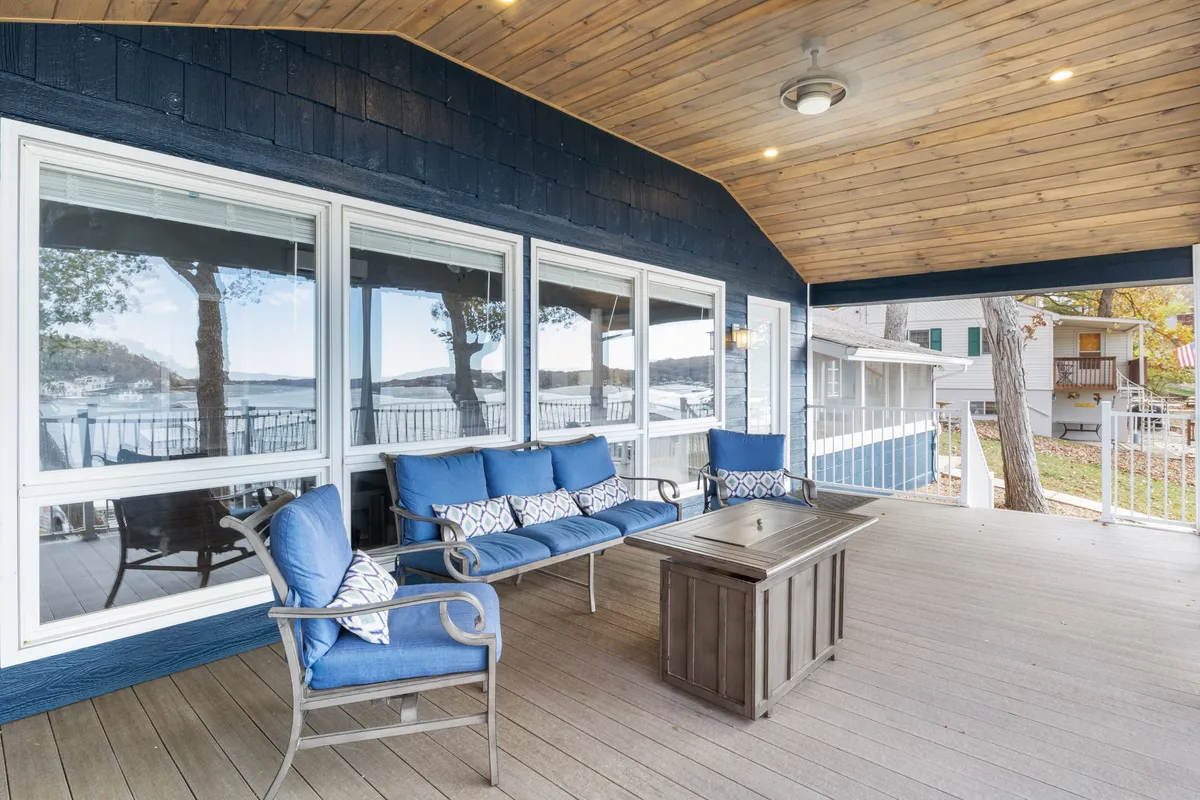 A cozy outdoor seating arrangement on a patio, with comfortable cushioned seats, decorative pillows, and large windows offering views of the lake. The wooden ceiling adds warmth, and the setup is ideal for casual relaxation or informal meals with family and friends.