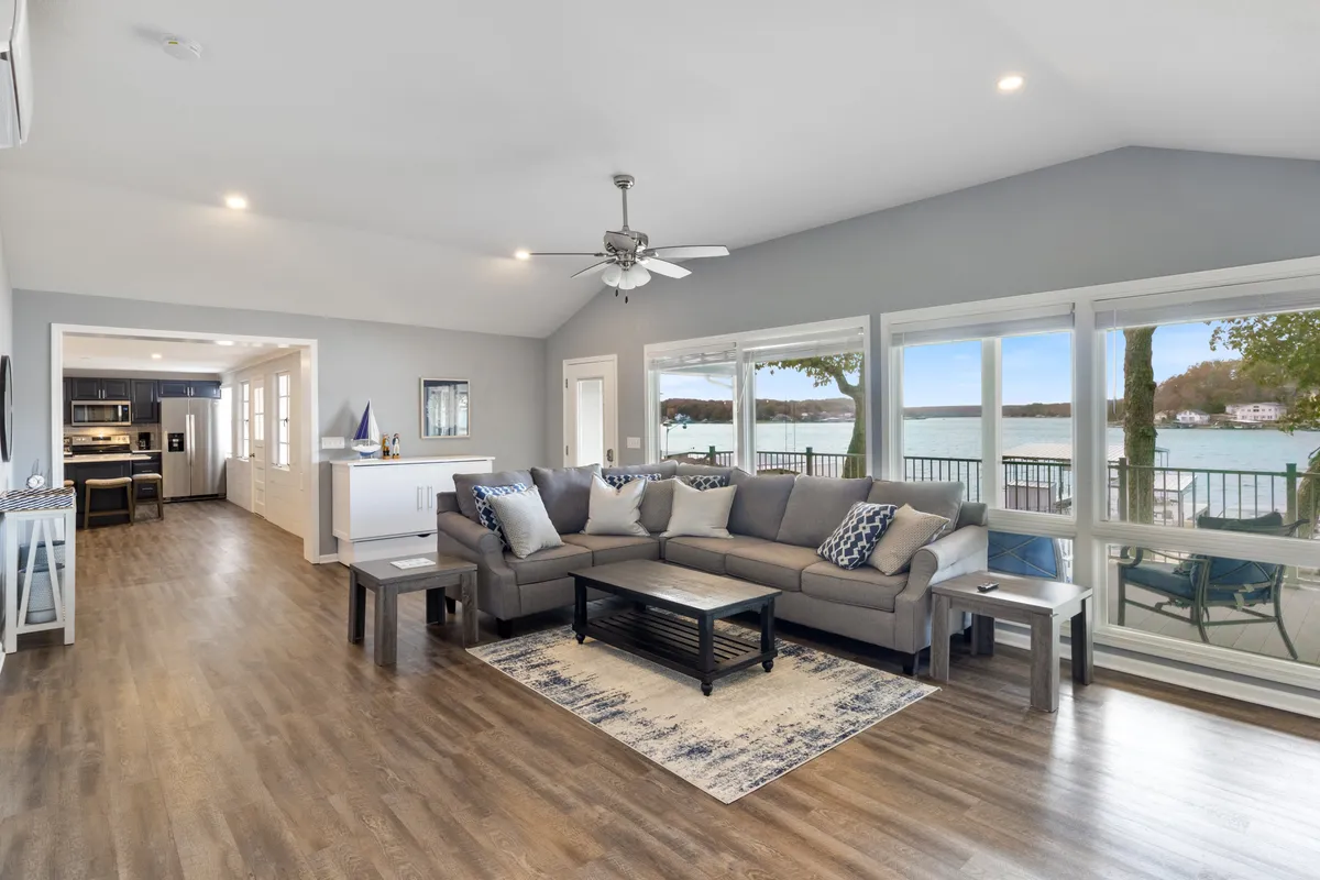 A spacious, open-plan living area featuring a large sectional sofa and coffee table, with expansive windows that offer stunning views of the lake. The arrangement welcomes gatherings and relaxation, while the natural light creates a warm, tranquil atmosphere.