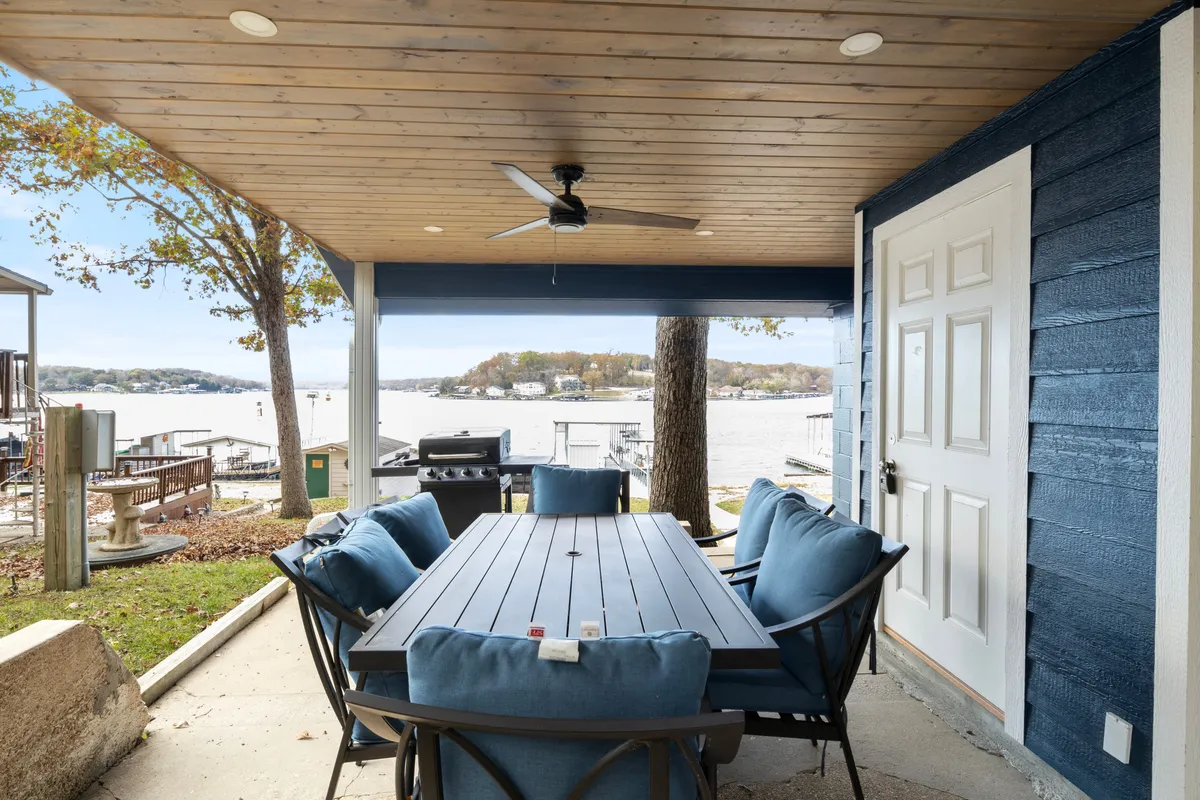 This image captures an outdoor dining table with blue chairs set on a patio overlooking the lake, with a grill nearby. The setup is clearly designed for al fresco dining, and the scene exudes a relaxed, vacation-like vibe with the shimmering water and lush greenery in the background.