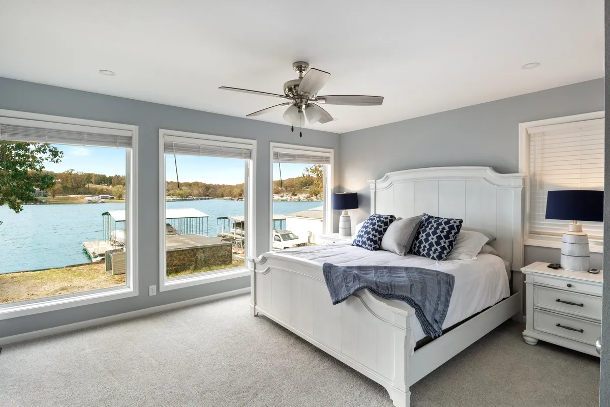 A serene bedroom featuring a large white bed with decorative pillows, a bedside table with a lamp, and expansive windows that open up to a beautiful view of the lake. The sunlight floods the space, and the calm atmosphere makes it ideal for relaxation or reading by the window.
