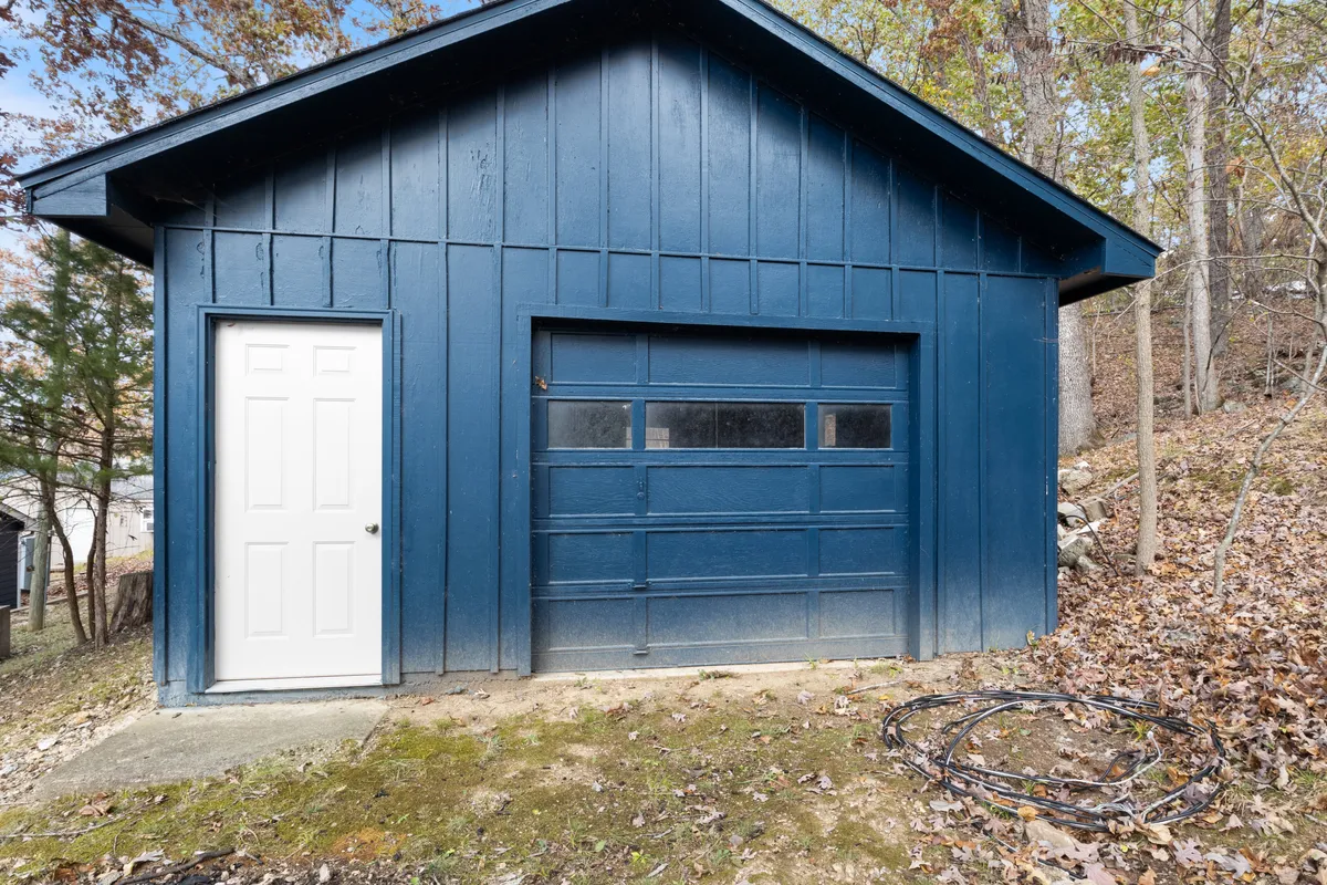 A blue garage with a white door is nestled among mature foliage, hinting at a practical base for spontaneous trips to the Lake of the Ozarks. The scene suggests easy access and readiness for exploration and adventure, with nature framing the structure.
