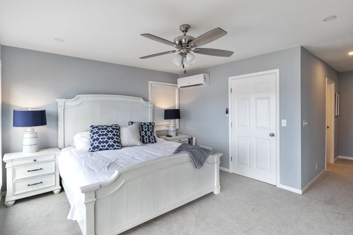 A cozy bedroom with a white bed frame, blue accents, nightstands with matching lamps, and decorative pillows. Sunlight streams into the space, creating a bright and inviting atmosphere that's perfect for relaxing or enjoying a restful night's sleep.